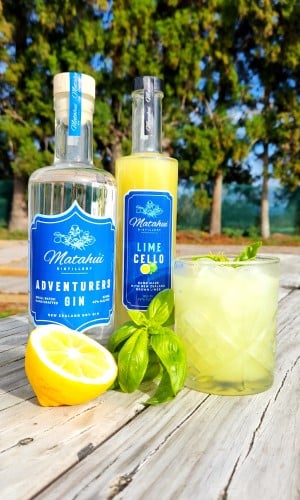 Delicious Basil Gin Sour cocktail served in an old fashioned glass with plenty of ice. Displayed on a wooden table with the ingredients: cut lemon, fresh basil sprig, Adventurers Gin and Matahui Limecello.